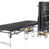 Escenario plegable móvil con ruedas para eventos, mostrando un módulo desplegado y varios paneles plegados en almacenamiento vertical.