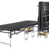 Escenario plegable móvil con ruedas para eventos, mostrando un módulo desplegado y varios paneles plegados en almacenamiento vertical.